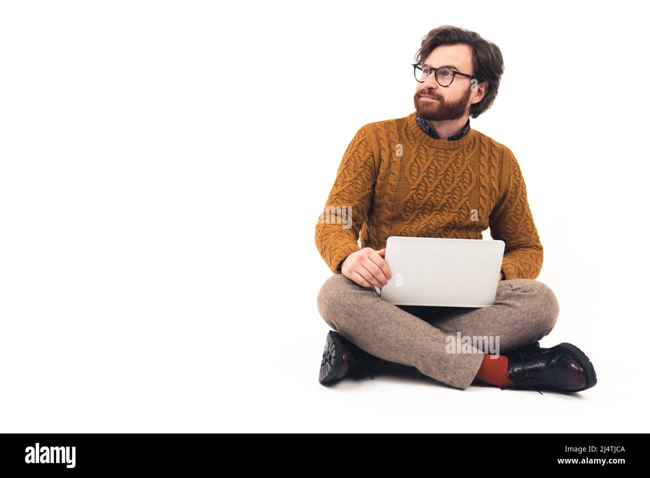 Bärtiger Mann mit moderner Brille, sitzend mit gekreuzten Beinen und Laptop auf den Knien haltend, der von der Kamera wegschaut. Hochwertige Fotos Stockfoto