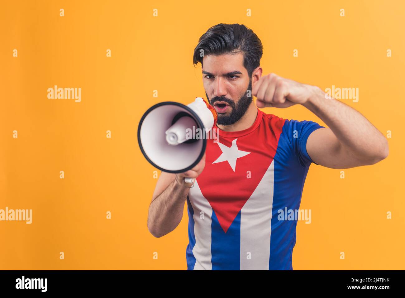 Junge kaukasische bärtige attraktive aktive Führer glücklichen Mann 20s trägt kubanische Flagge T-Shirt sprechen in Lautsprecher isoliert auf gelbem Hintergrund Studio-Porträt. Hochwertige Fotos Stockfoto