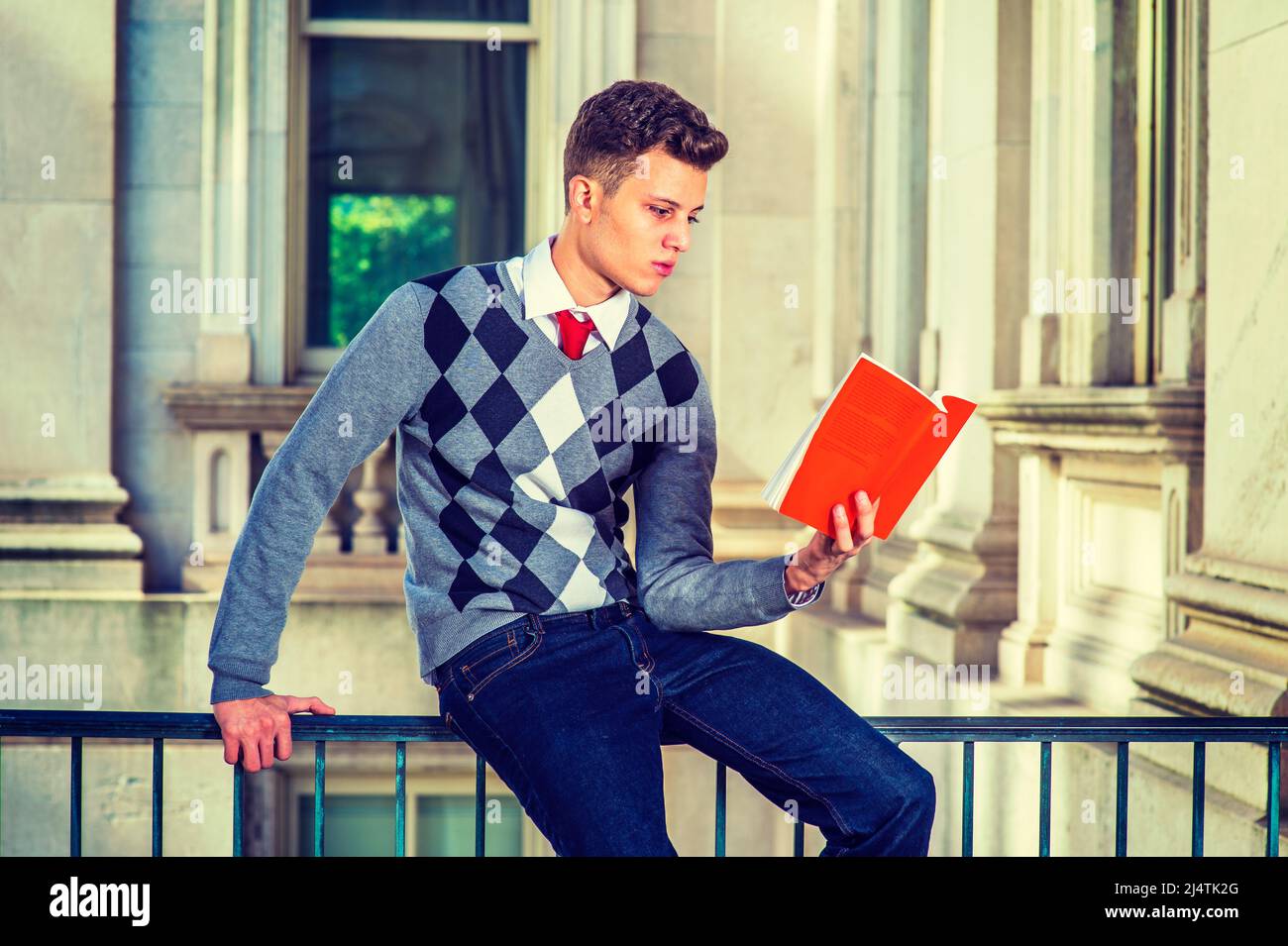 Mann Beim Lesen Draußen. In einem schwarzen, weißen, grau gemusterten Pullover, Jeans, sitzt ein junger, hübscher Student auf einem Geländer im Büro b Stockfoto