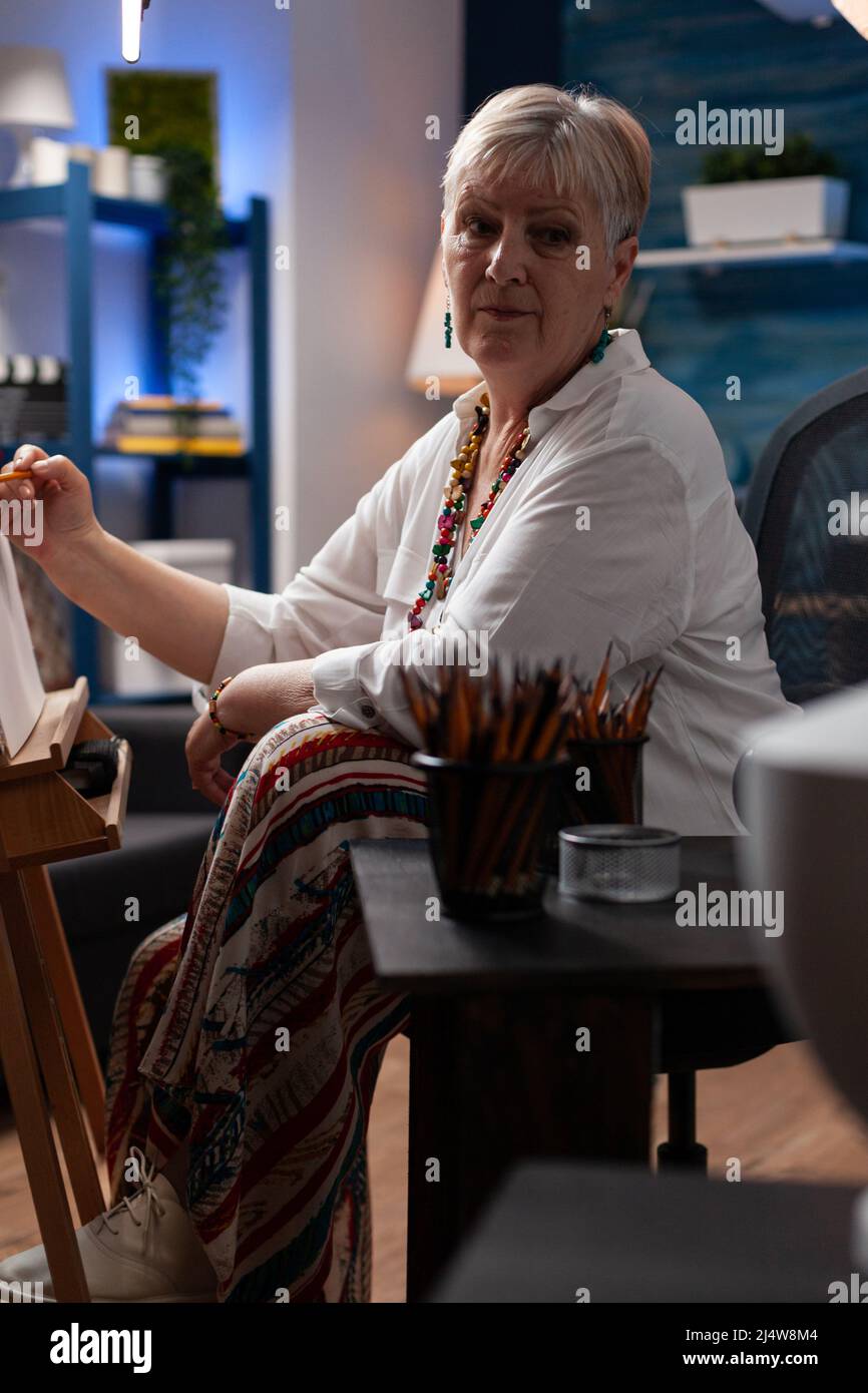 Porträt von grauhaarigen älteren Hobby-Künstler Studium Schattierung Licht auf Stillleben mit Modell Vase in Home Studio. Pensionierte Frau zeichnet mit hb-Bleistift auf Leinwand vor der Staffelei, während sie sich hinsetzte. Stockfoto