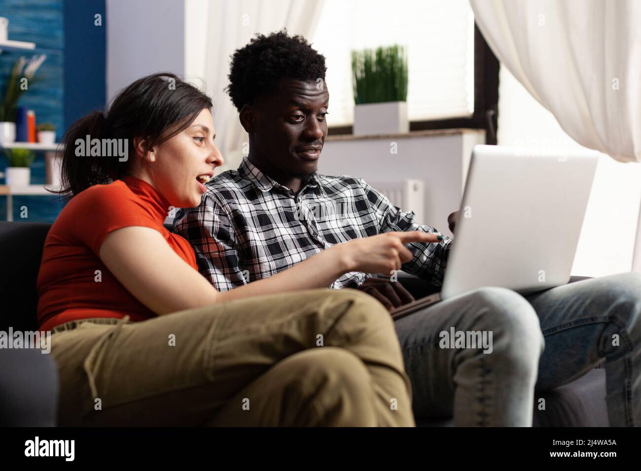 Fröhliches junges multiethnisches Paar, das auf den Computerbildschirm schaut, während es auf der Couch im Wohnzimmer sitzt. Menschen genießen es, beim lässigen Surfen zusammen zu sein und sehen interessante Videos im Internet. Stockfoto