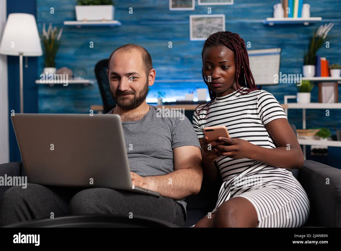 Glücklicher kaukasischer Mann websurfing auf Laptop, während afroamerikanische Frau Smartphone hält und auf den Computerbildschirm schaut. Ein Paar sitzt auf dem Sofa im Wohnzimmer und überprüft Rechnungen und finanzielle Probleme am Computer und Telefon Stockfoto
