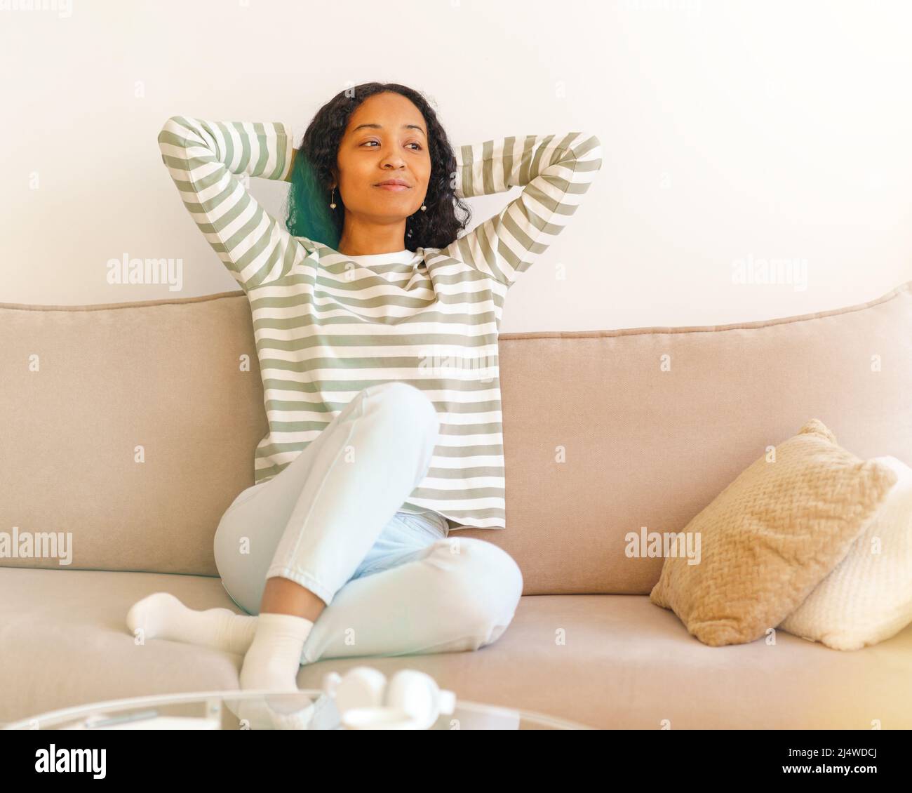 Lächelnde afroamerikanische Frau, die die Hände hinter den Kopf legt, während sie auf dem Sofa im Wohnzimmer sitzt Stockfoto