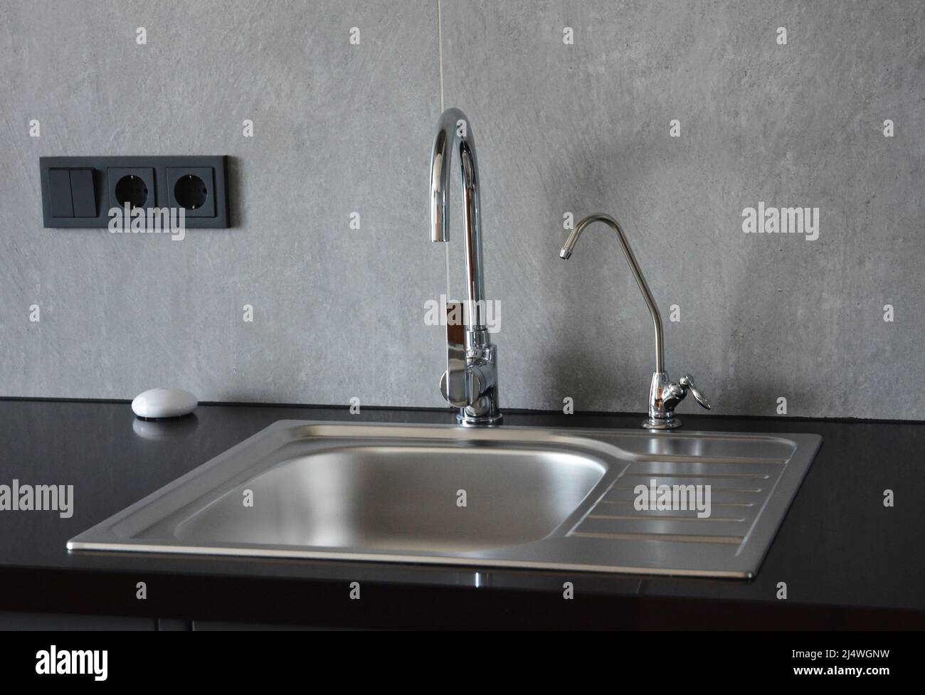 Ein Küchenspüle aus Metall auf einer schwarzen Küchenarbeitsfläche mit zwei Wasserhähnen, wobei einer ein kleiner Filterhahn für Trinkwasser ist. Stockfoto