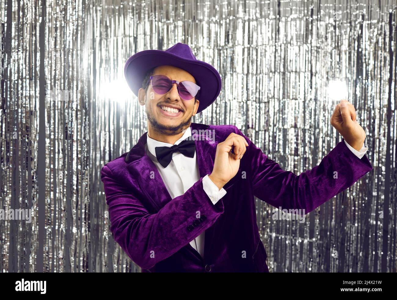 Fröhlicher, lustiger junger Mann mit violetter Jacke, Hut und Sonnenbrille, der auf der Party tanzt Stockfoto
