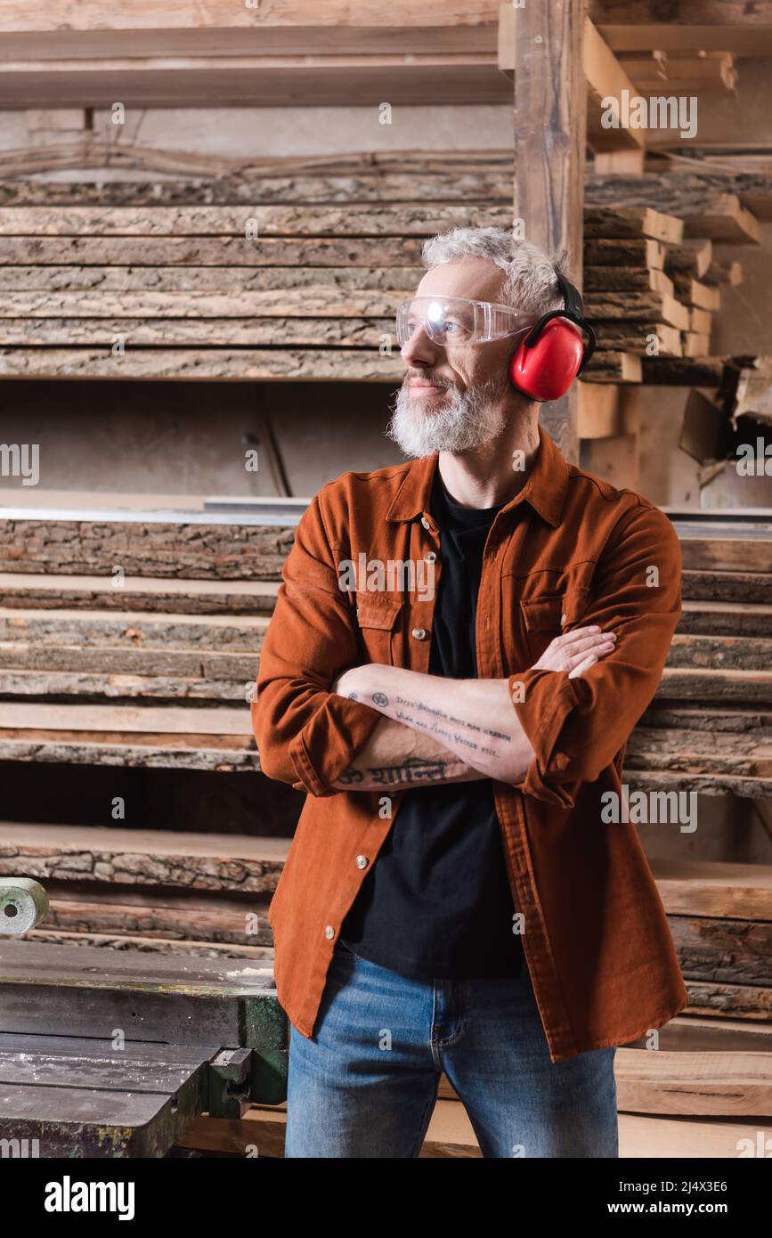 Bärtiger Möbeldesigner, der mit gekreuzten Armen in der Nähe von Brettern in der Werkstatt steht Stockfoto