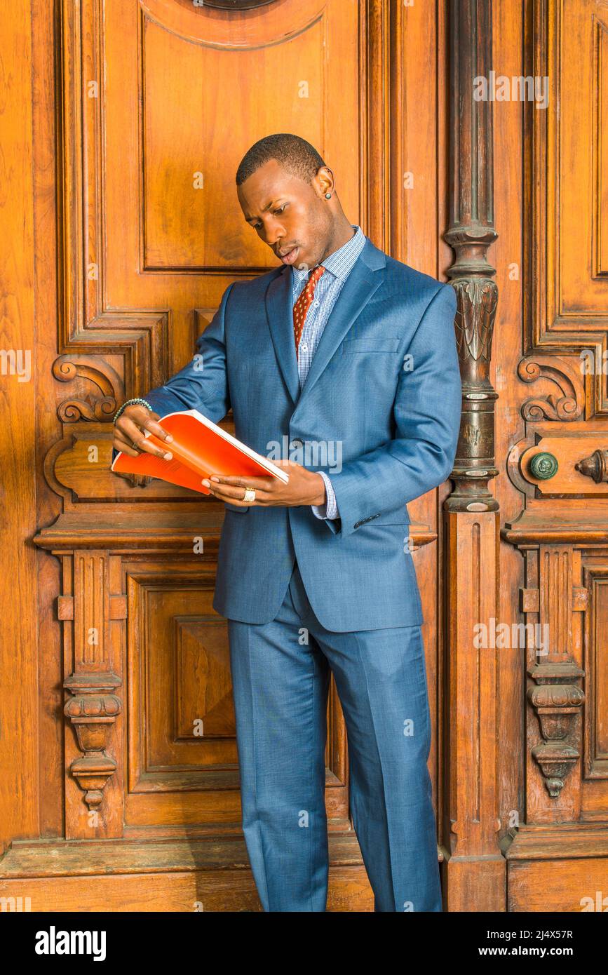 Formell in blauem Anzug, gemustertes Hemd, Krawatte, kurzer Haarschnitt, Ohrstecker tragen, Eine junge schwarze Lehrerin steht vor einem alten Stil Stockfoto