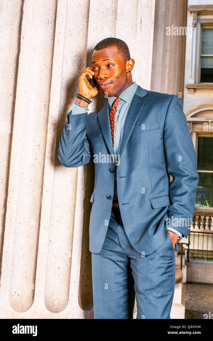 Amüsiert durch Telefonanruf. In blauem Anzug, Krawatte, ein junger schwarzer Geschäftsmann, der vor dem Büro an einer Säule stand, die Augenbrauen hob, die Augen öffnete und Li schloss Stockfoto