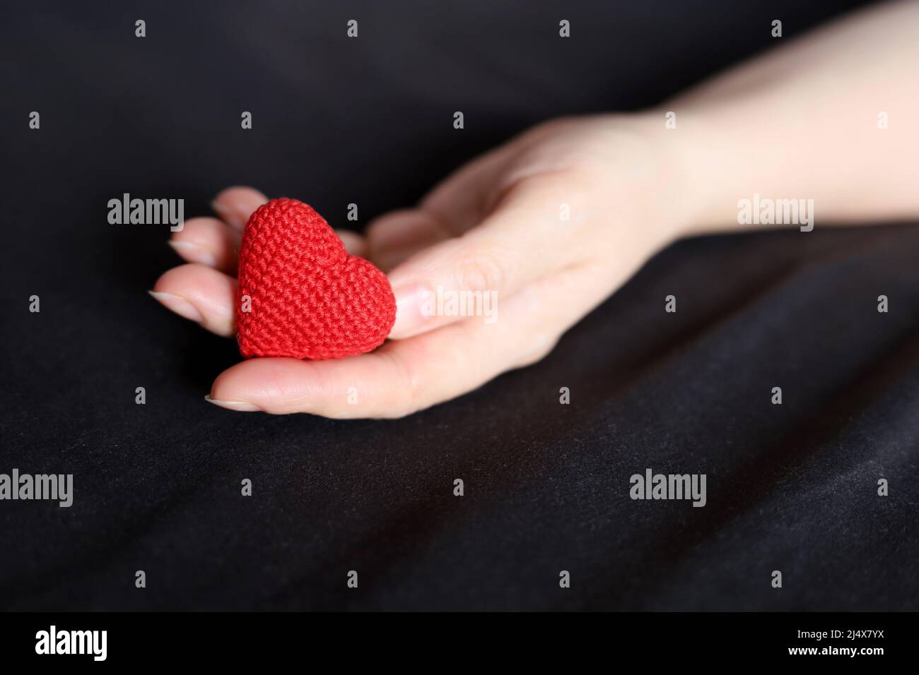 Rot gestricktes Herz in weiblicher Handfläche auf einem Bett mit schwarzer Bettwäsche. Konzept der Liebe, Gesundheitsversorgung, Mutterschaft, Blutspende Stockfoto