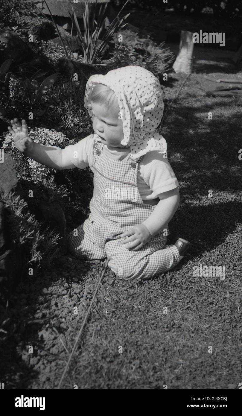 1960s, historisch, ein Kleinkind mit einer gepunkteten Baumwollkopfhaube, draußen, sitzend auf den Knien bei einem Blumenbett, England, Großbritannien. Stockfoto