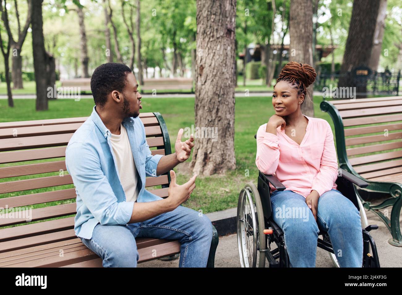 Junger afroamerikanischer Mann, der auf der Bank sitzt und mit seiner Freundin mit körperlicher Behinderung im Freien spricht Stockfoto