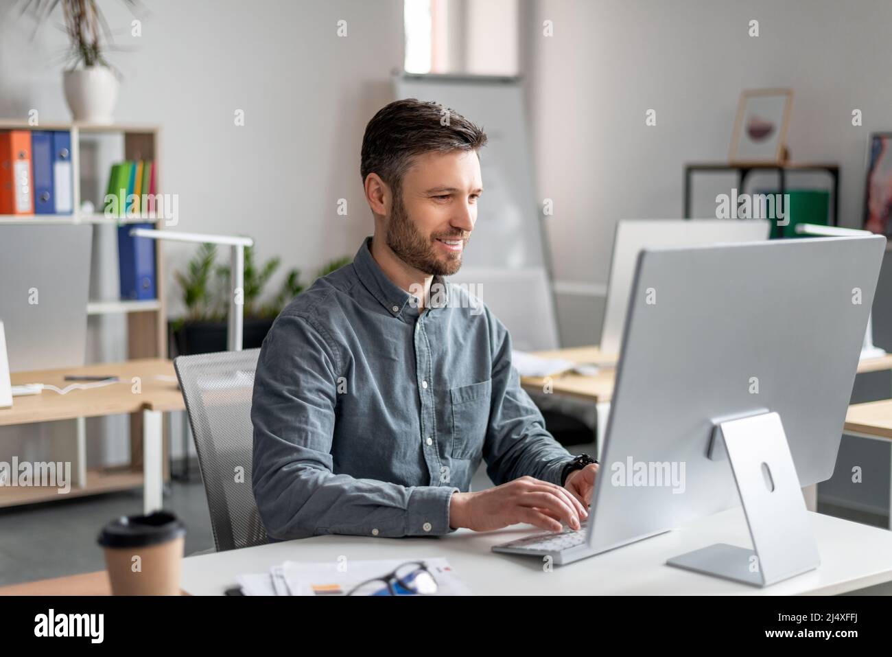 Fröhlicher Geschäftsmann mittleren Alters, der am Arbeitstisch sitzt, auf der Computertastatur tippt und an Marketingforschung arbeitet Stockfoto