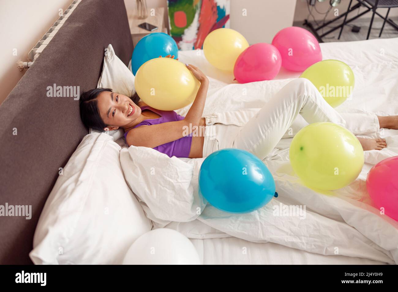 Fröhliche asiatische Frau, die im Bett mit bunten Ballons liegt Stockfoto