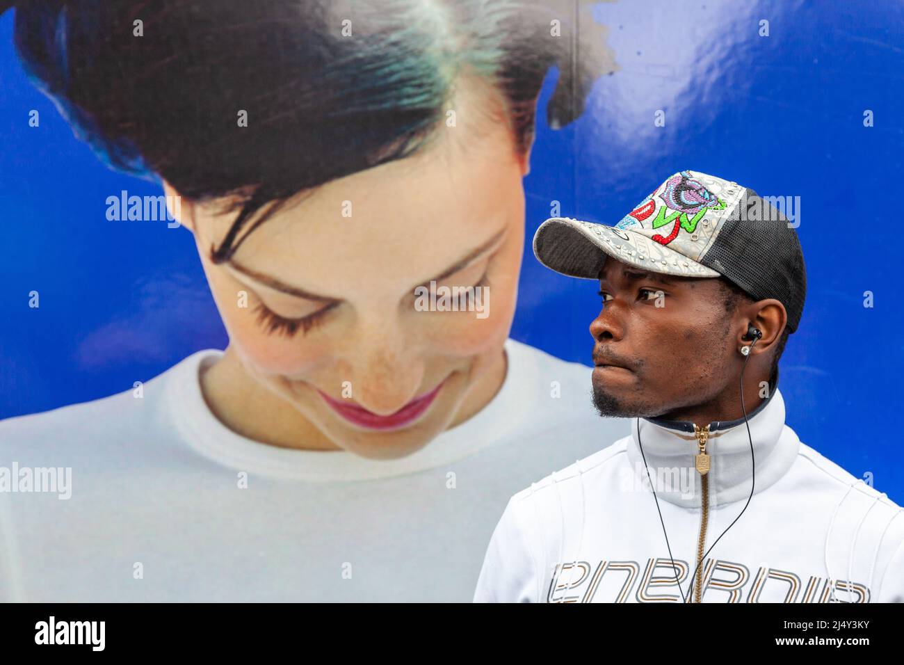 Ein eingeklemmter Mann mit einer Spitzenmütze steht vor einem riesigen Foto einer jungen Frau, die lächelnd herunterschaut. Brüssel. Stockfoto