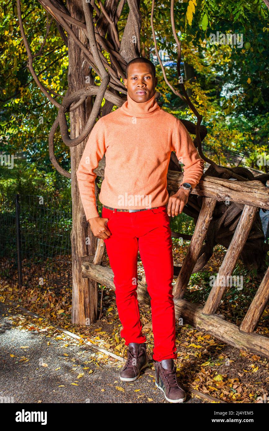 Männer Herbst Casual Fashion. Kleid in hellorange Pullover mit hohem Kragen, rote Hosen, gemusterte Stiefel Schuhe, Tragen Armbanduhr, ein junger schwarzer Kerl i Stockfoto