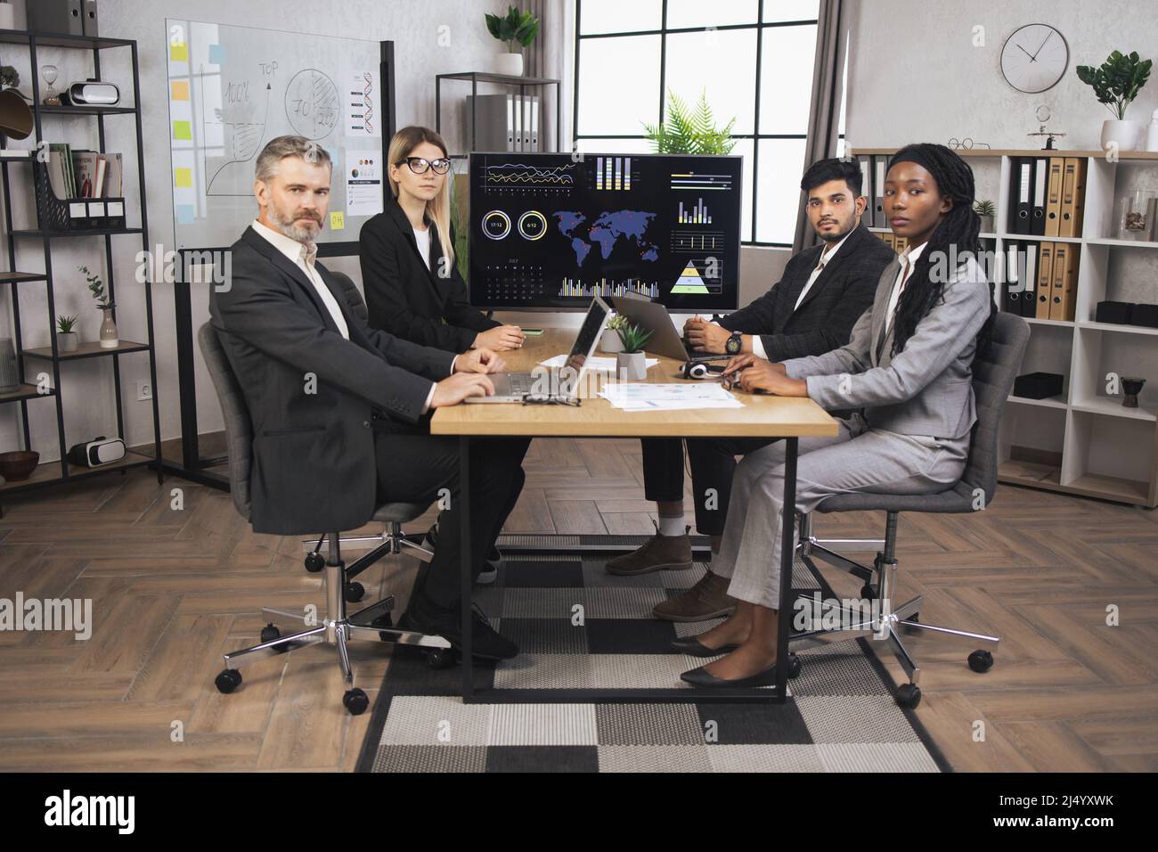 Das Team aus vier verschiedenen multiethnischen Geschäftsleuten trifft sich im Sitzungssaal des Innenbüros, sitzt mit Tablets und Laptop am Schreibtisch, vor einem riesigen Plasma-TV-Bildschirm und blickt auf die Kamera. Stockfoto