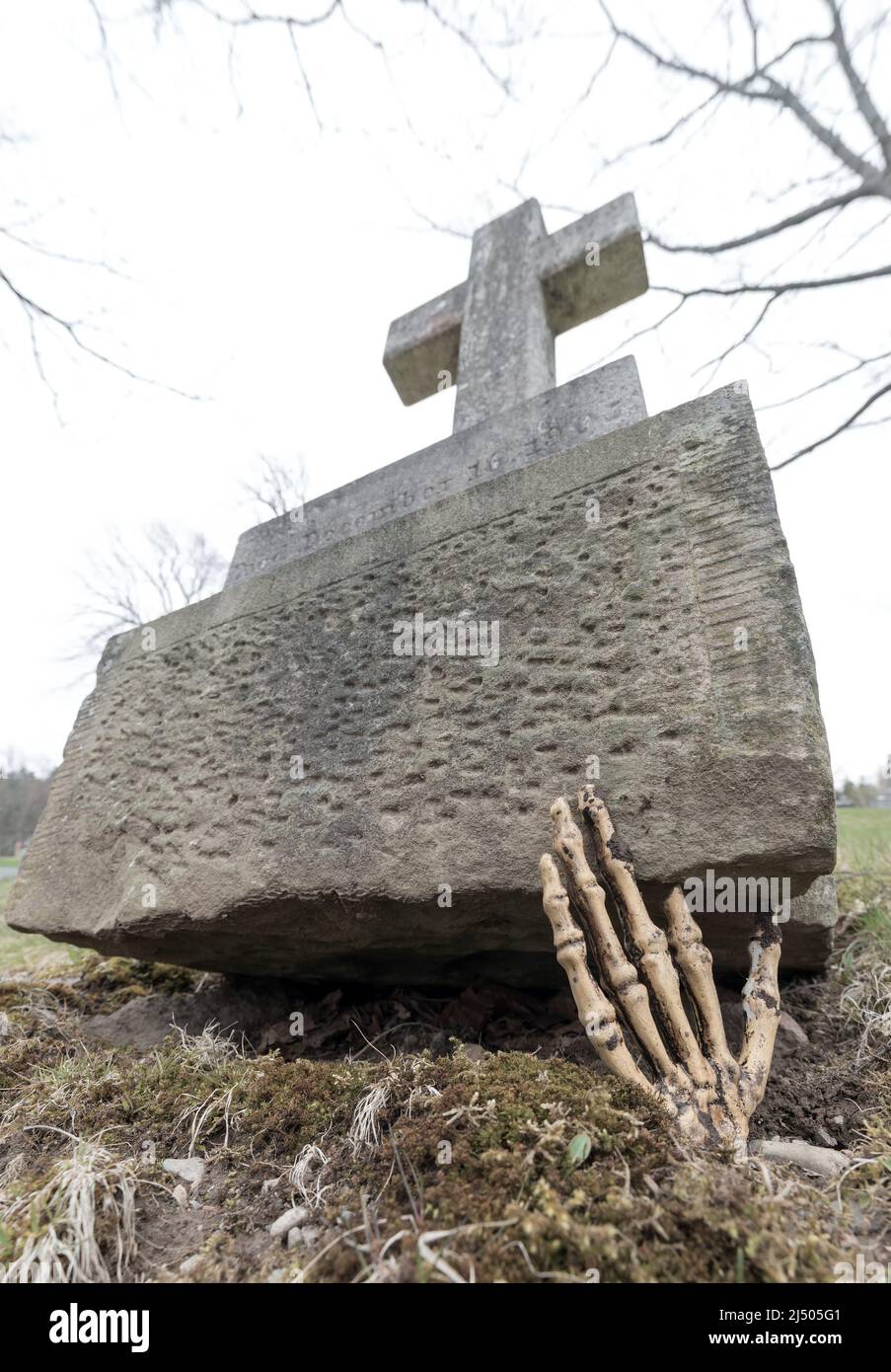 Eine skelettartige Hand, die aus dem Untergrund greift, um ein Grab hinüber zu schieben. Die Hand ist mit Schlamm verschmutzt. Es gibt keinen Namen auf dem Grab, aber ein Datum ist lesbar. Stockfoto