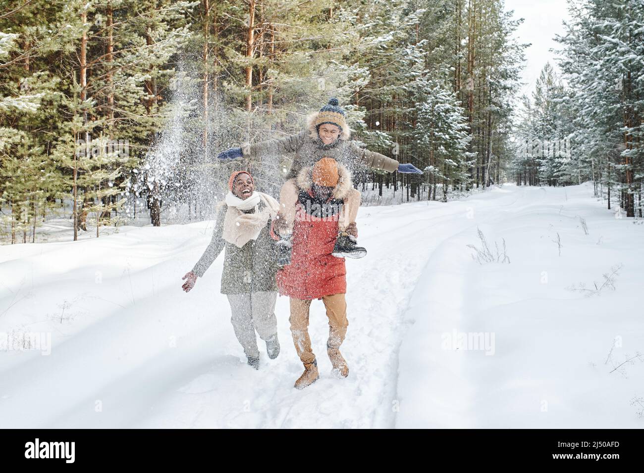 Fröhliche afroamerikanische Familie in Winterkleidung, die sich am verschneiten Tag amüsieren und Zeit im Park oder Wald zwischen immergrünen Bäumen verbringen Stockfoto