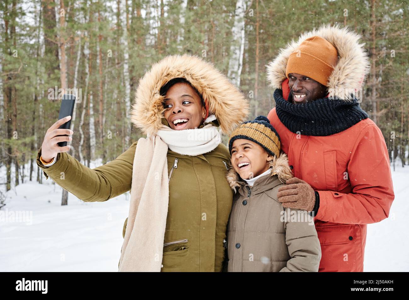 Glückliche, liebevolle afroamerikanische Familie in warmer Winterkleidung, die Selfie im Park oder Wald macht, während die Frau das Smartphone hält Stockfoto