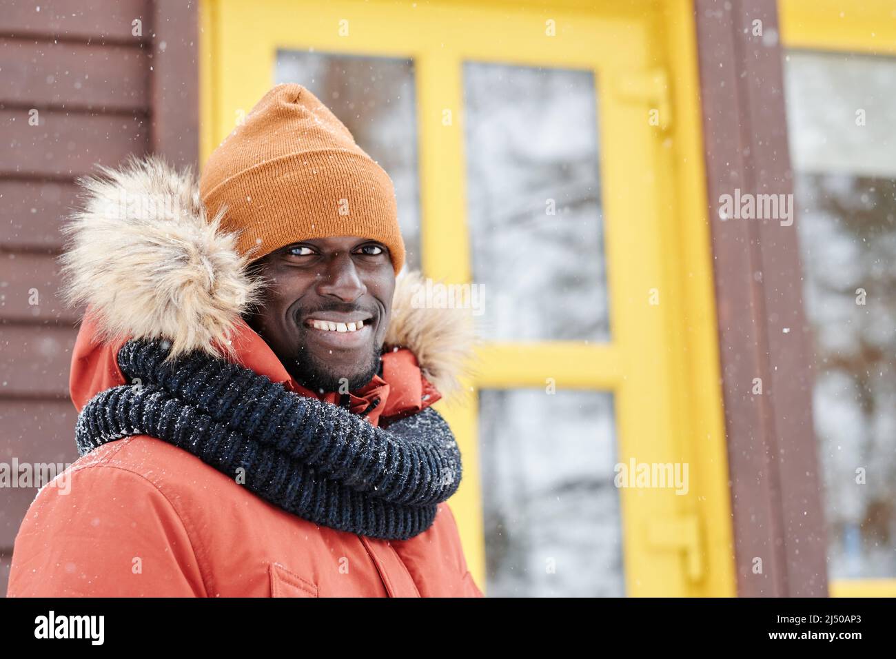 Junger lächelnder schwarzer Mann in Winterjacke, Mütze und warmem Wollschal, der vor der Kamera gegen das neue Landhaus steht Stockfoto