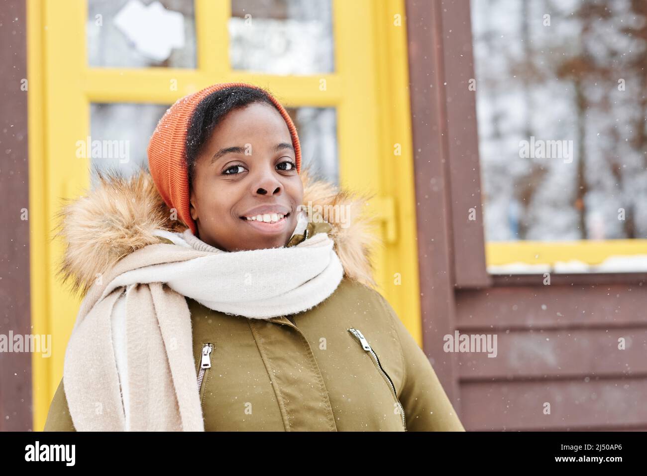 Fröhliche afroamerikanische Frau in Winterjacke, Mütze und weißem Wollschal, die vor der Kamera gegen das neue Landhaus schaut Stockfoto