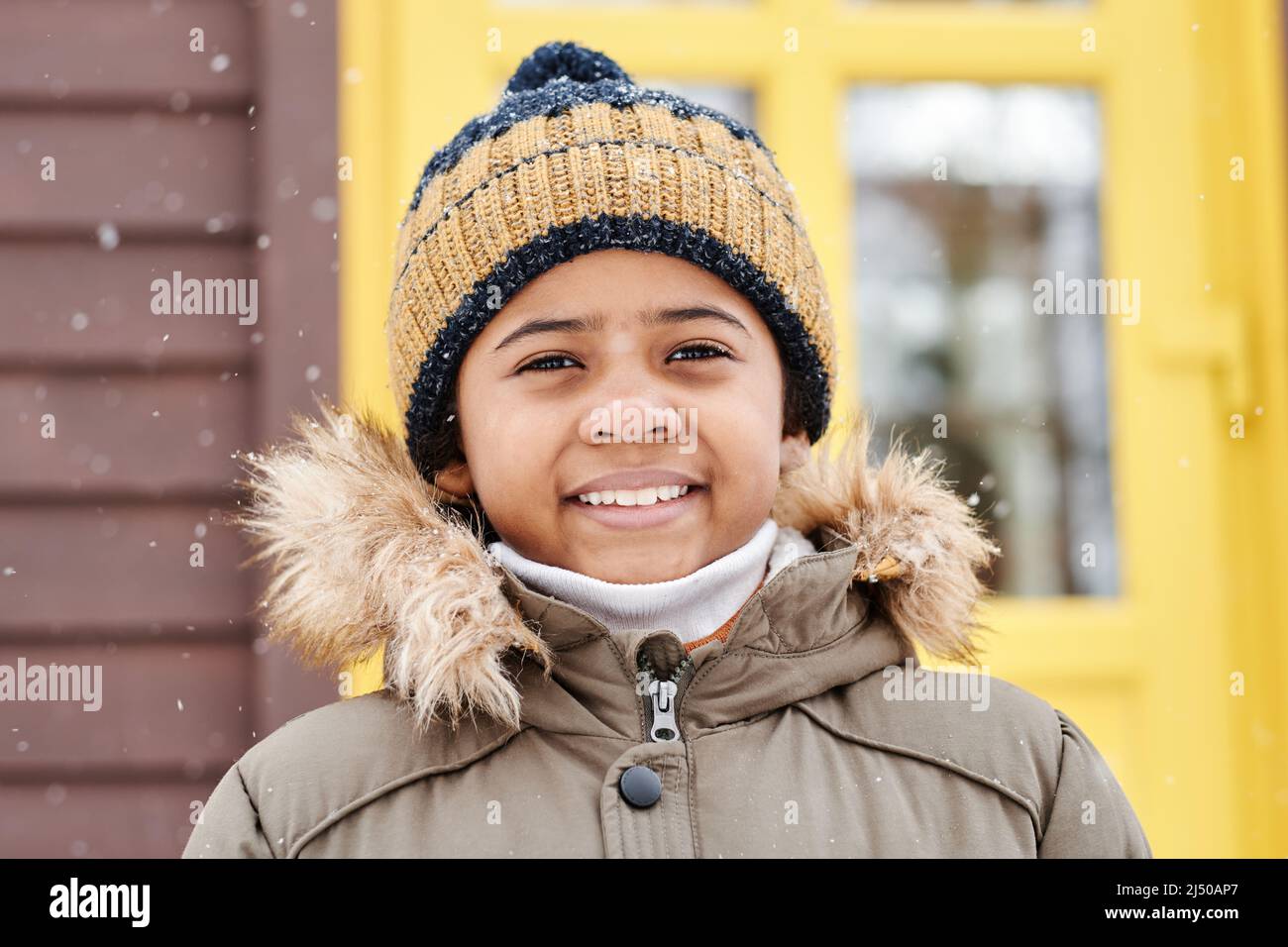 Happu entzückender Junge in warmer Winterjacke und gestreiftem Strickmütze, der die Kamera anschaut, während er gegen die gelbe Tür des Landhauses steht Stockfoto