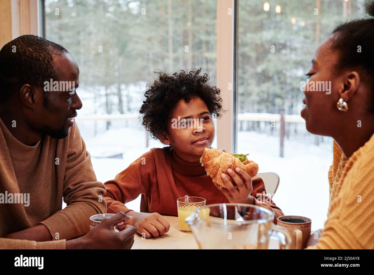 Süßer afroamerikanischer Junge mit hausgemachtem Hotfog, der beim Frühstück seiner Mutter zuhört, während eine dreiköpfige Familie am Tisch vor einem großen Fenster sitzt Stockfoto
