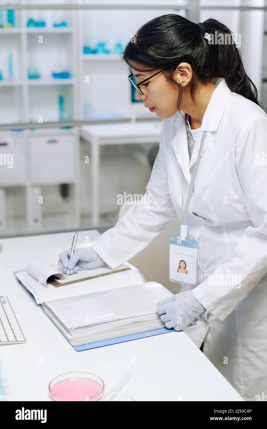 Junge Biochemikerin in Weißkochweiß und Handschuhe, die im Notizblock wissenschaftliche Notizen machen, während sie im klinischen Labor am Schreibtisch steht Stockfoto