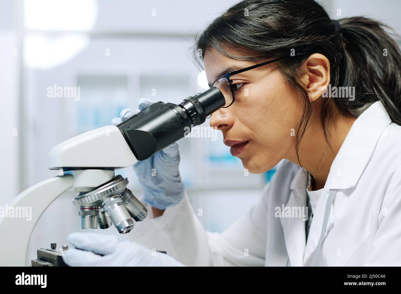 Seitenansicht einer jungen ernsthaften Biochemikerin in Weißkohlegrän, Handschuhen und Brillen, die während der wissenschaftlichen Forschung im Mikroskop betrachtet wurden Stockfoto