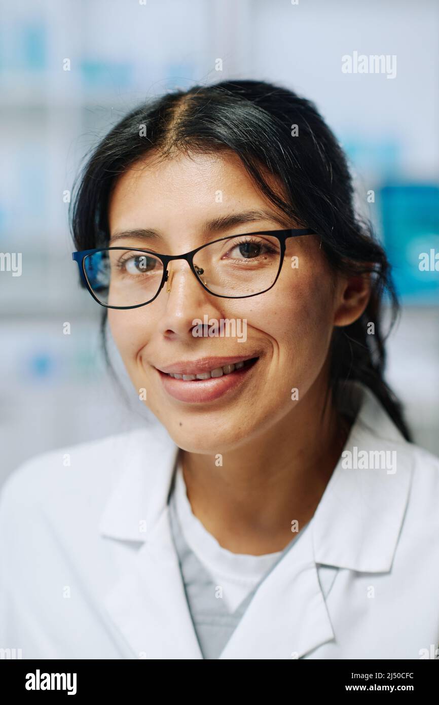 Junge, erfolgreiche hispanische Biochemikerin in Brillen und Weißkohlegräsen, die während ihrer Arbeit im Labor mit einem Lächeln auf die Kamera blickt Stockfoto