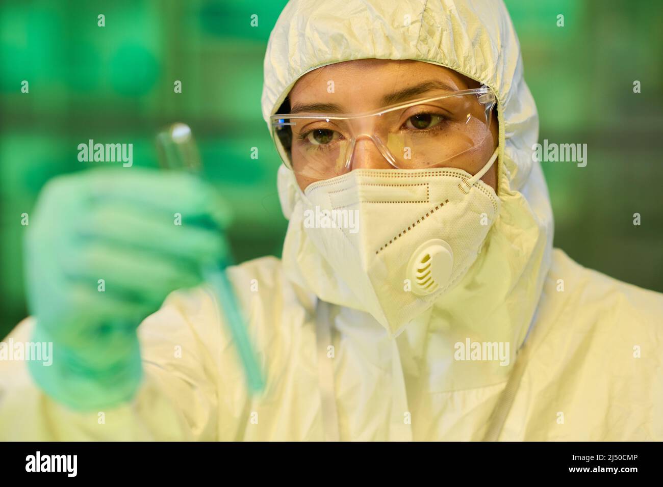 Eine Wissenschaftlerin in Schutzbrille, Atemschutzmaske und Biogefährdungsanzug, die chemische Proben in der Reagenzglas-Hand in ihrer Handschuhen ansieht Stockfoto