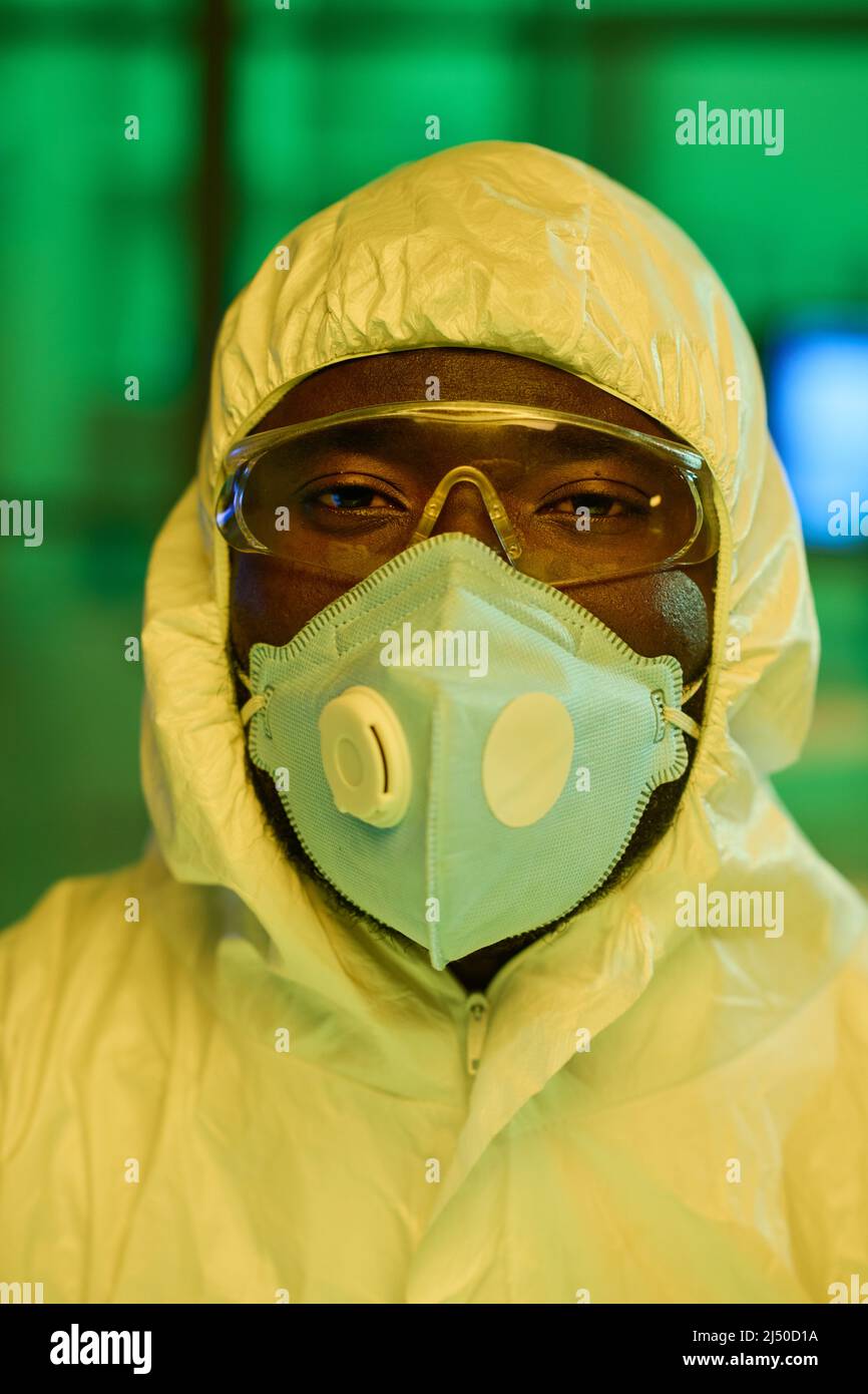 Zeitgenössischer schwarzer Mann in weißen Schutzhüllen, Atemschutzmaske und Brillen, der während der Arbeit im klinischen Labor auf die Kamera blickt Stockfoto