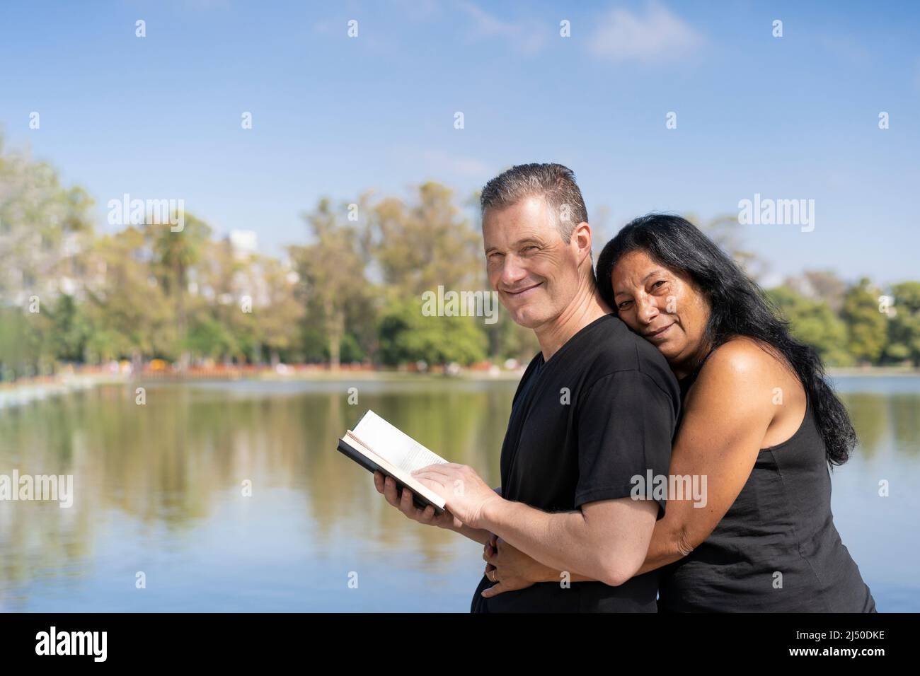 Reifes multiethnisches Paar, das sich an einem See umarmt und ein Buch liest und lächelnd die Kamera anschaut. Speicherplatz kopieren Stockfoto