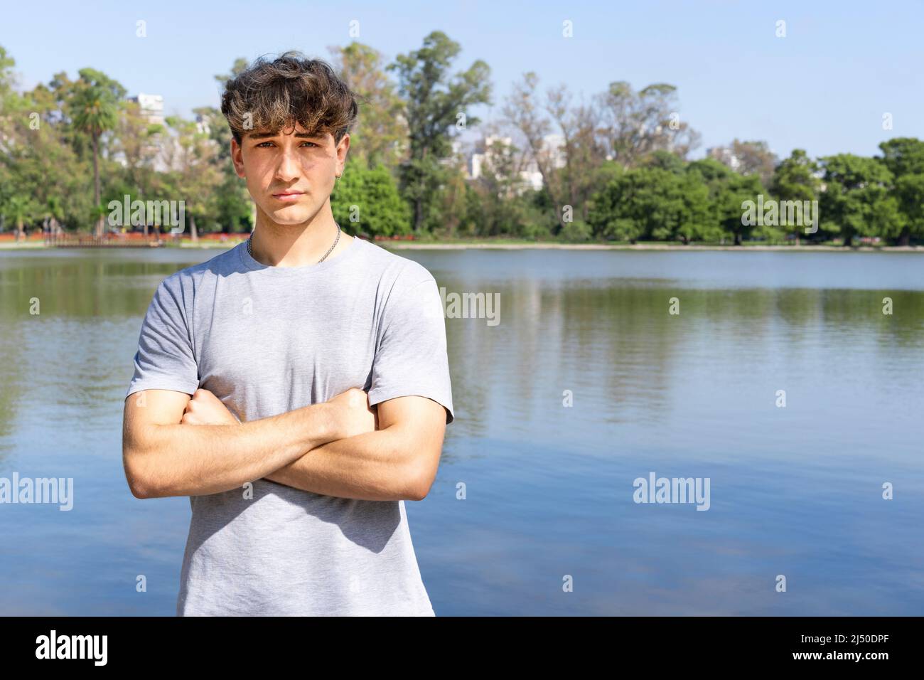 Porträt eines selbstbewussten jungen blonden Mannes mit gekreuzten Armen, der auf die Kamera schaut, im Hintergrund des Sommersees. Speicherplatz kopieren Stockfoto