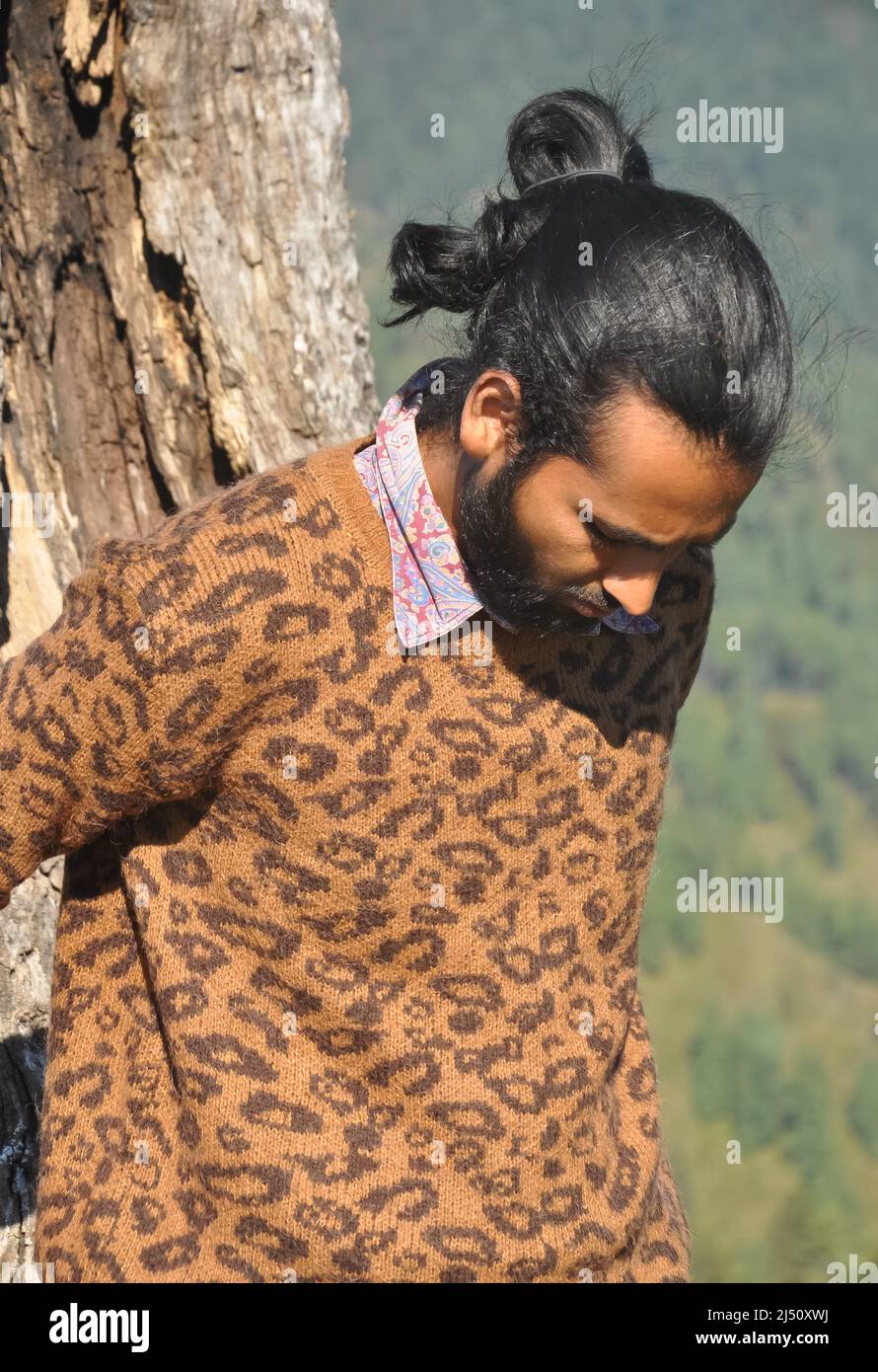 Nahaufnahme eines gut aussehenden indischen jungen Mannes mit einem Haarbüschen, das nach unten schaut, während er sich an einen Baum lehnt Stockfoto