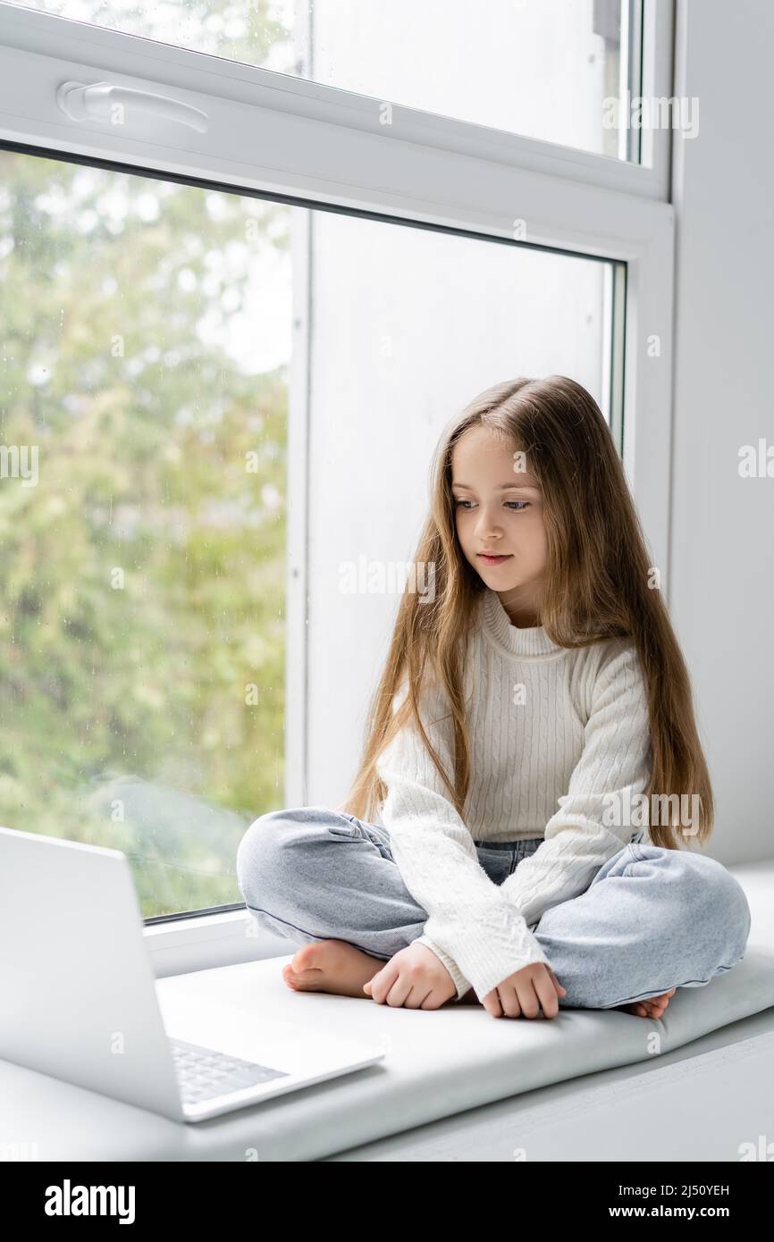 Barfuß Mädchen mit langen Haaren sitzt auf Fensterbank neben Laptop Stockfoto