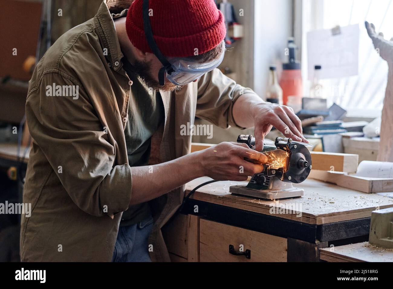 Professioneller junger männlicher Tischler mit Schutzbrille, der in der Schreinerei arbeitet und Messer mit Schleifvorrichtung schärft Stockfoto