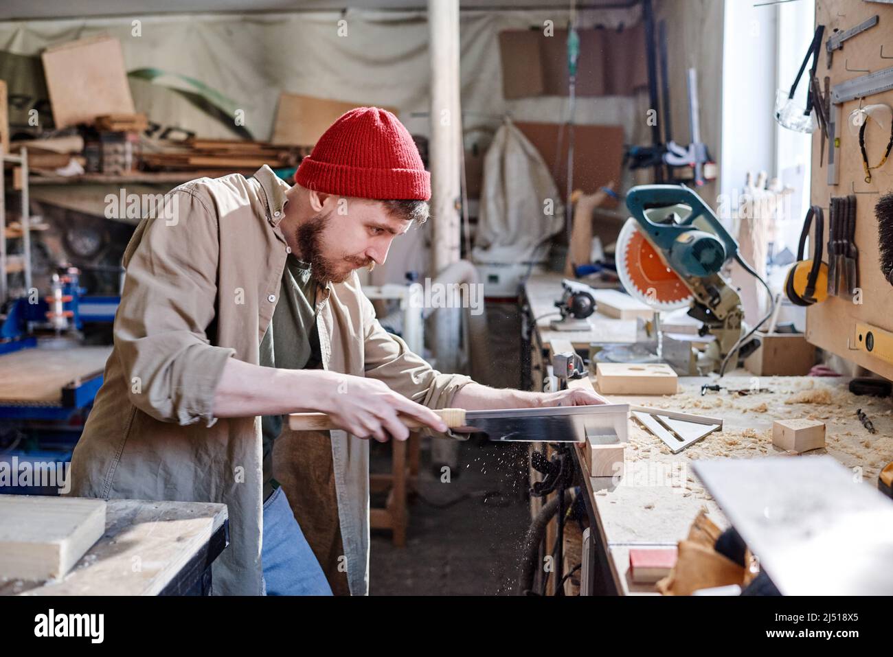 Horizontale Seitenansicht des jungen professionellen Zimmermanns, der Holzdiele mit Handsäge in der Holzwerkstatt schneidet Stockfoto
