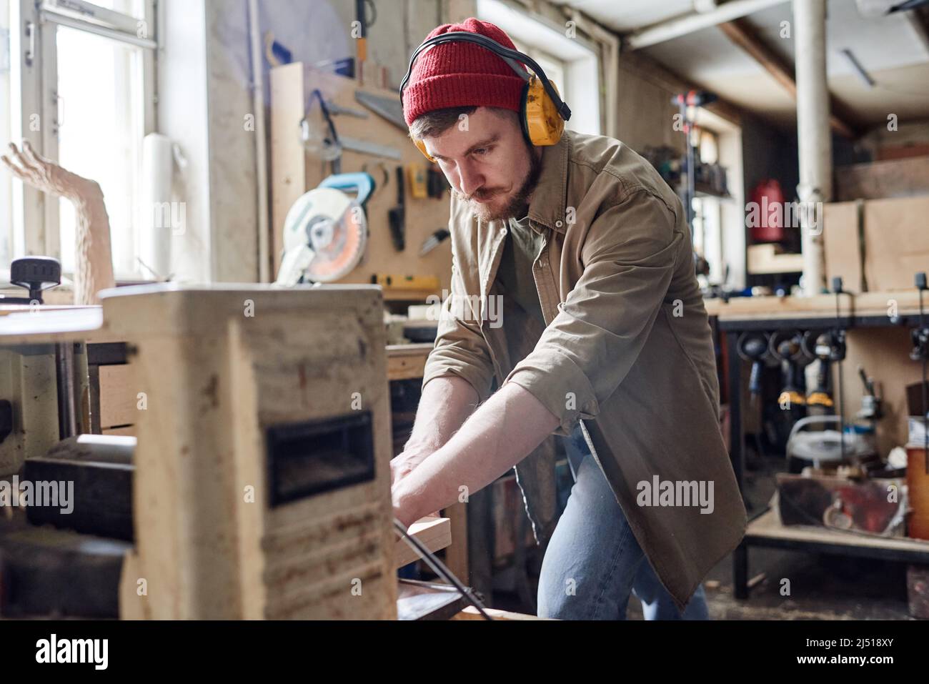 Junger Zimmermann mit geräuschreduzierenden Kopfhörern mit Dickenhobelmaschine in der Holzwerkstatt Stockfoto