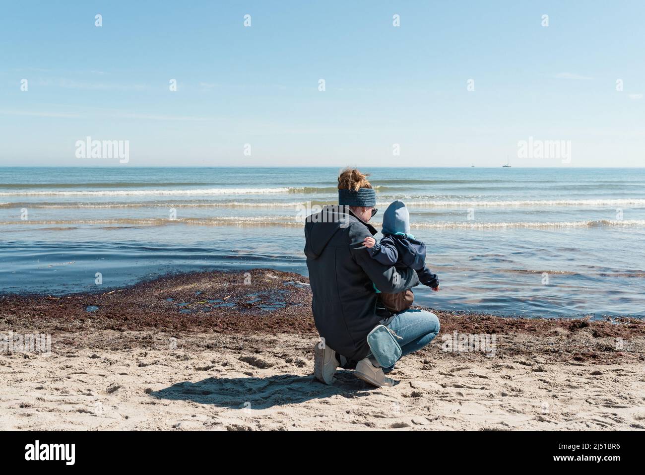 Rückansicht der Mutter, die ein Kleinkind am Strand hält, gegen das blaue Meer und den klaren blauen Himmel Stockfoto