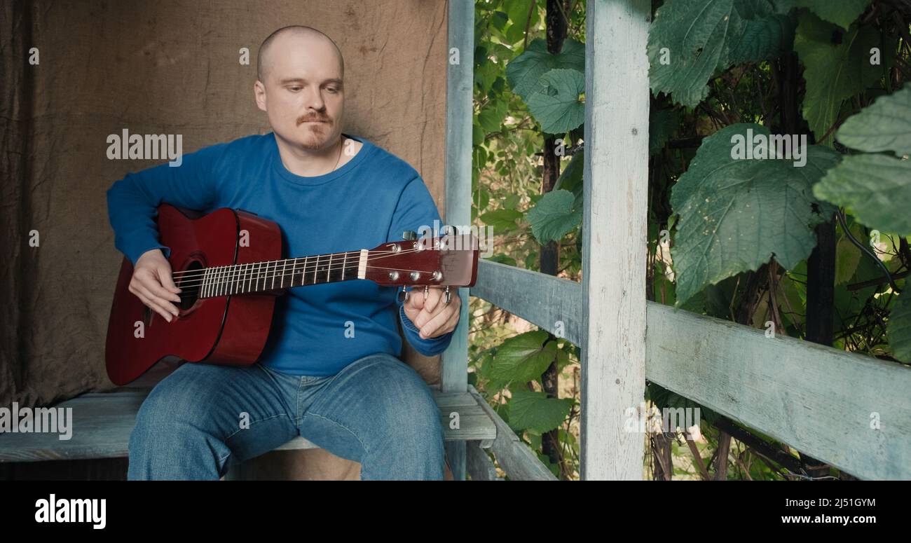 Mann mit einer akustischen Gitarre sitzt auf der Veranda des Bauernhauses, er stimmt das Instrument Stockfoto