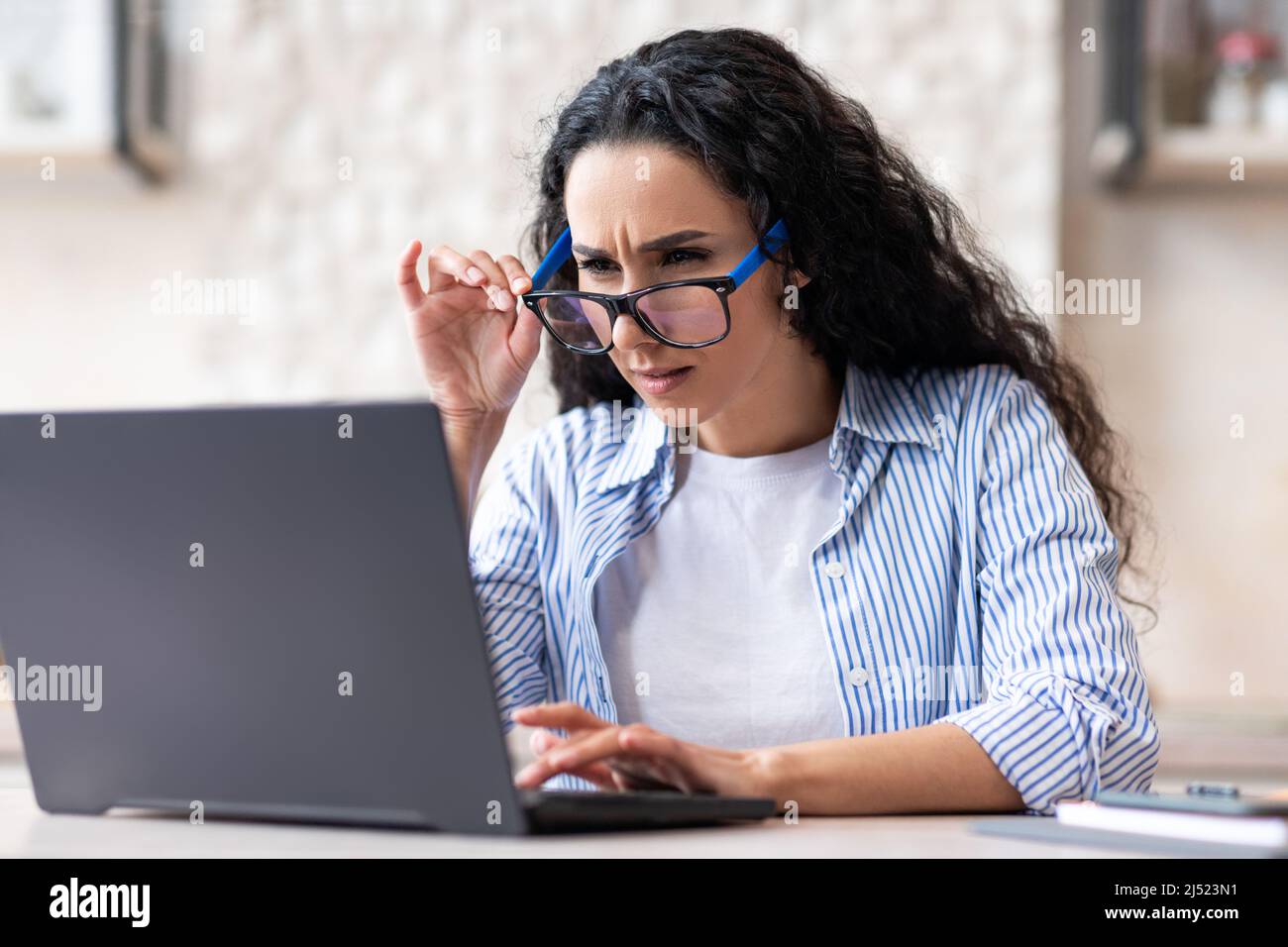 Sehstörungen. Eine lateinische Dame benutzt eine Brille, während sie zu Hause am Laptop arbeitet und in der Küche sitzt Stockfoto