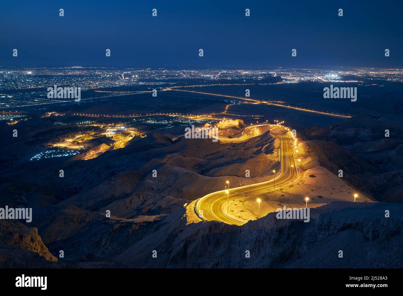 Kurvenreiche Straße in den Bergen gegen die beleuchtete Stadt bei Nacht. Al Ain im Emirat Abu Dhabi. Stockfoto