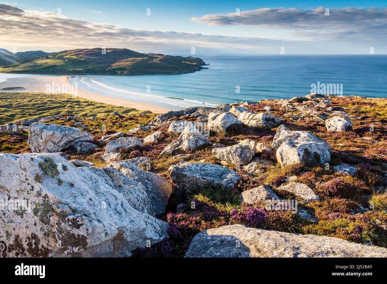 Ein Hügel mit Felsen und Heidekraut bietet einen wunderschönen Blick auf den einsamen Sandstrand und das herrlich türkisfarbene Meer der Torrisdale Bay, das in Sonnenschein getaucht ist. Stockfoto