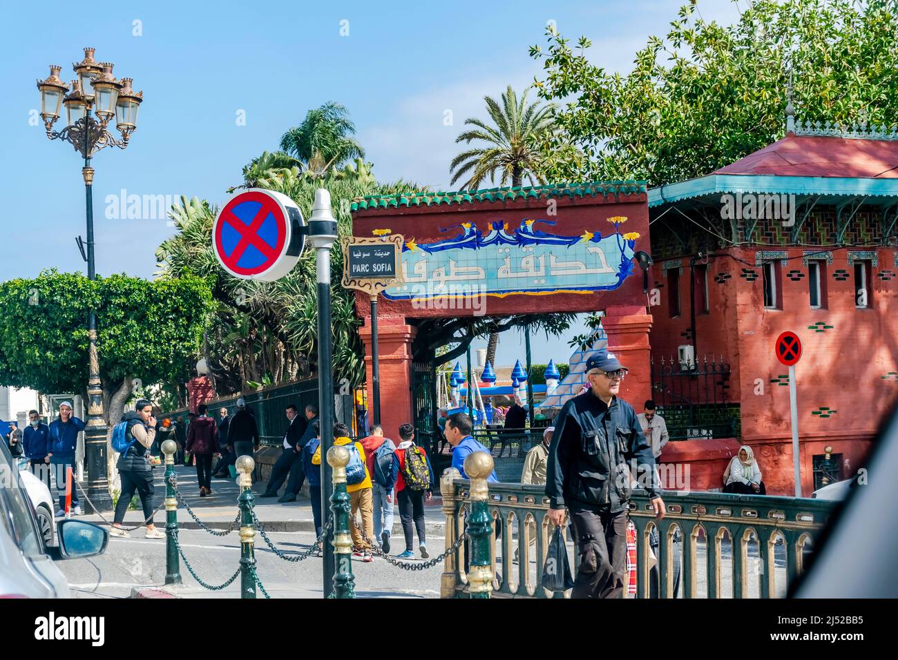 Sofia-Park-Eintrag mit einem Namensschild in arabischen Zeichen und französischer Sprache. Eine Menge Leute, die gehen und sitzen, Palmen und Lichtmast. Stockfoto