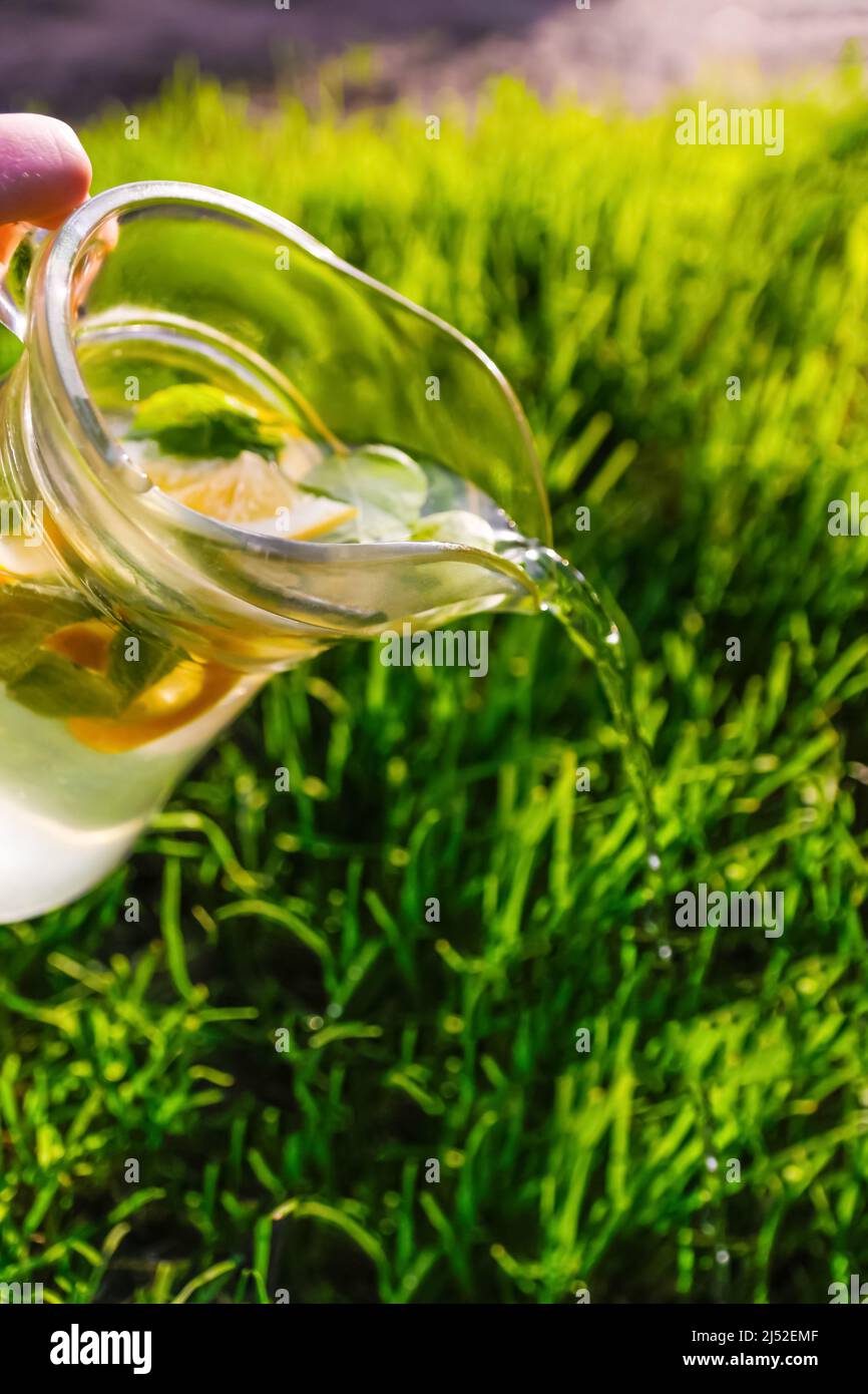 Unschärfe-Hand hält Glaskrug Limonade mit Zitrone und Minze auf natürlichem grünen Hintergrund. Krug mit einem kühlen Sommercocktail oder Punsch im Freien Stockfoto