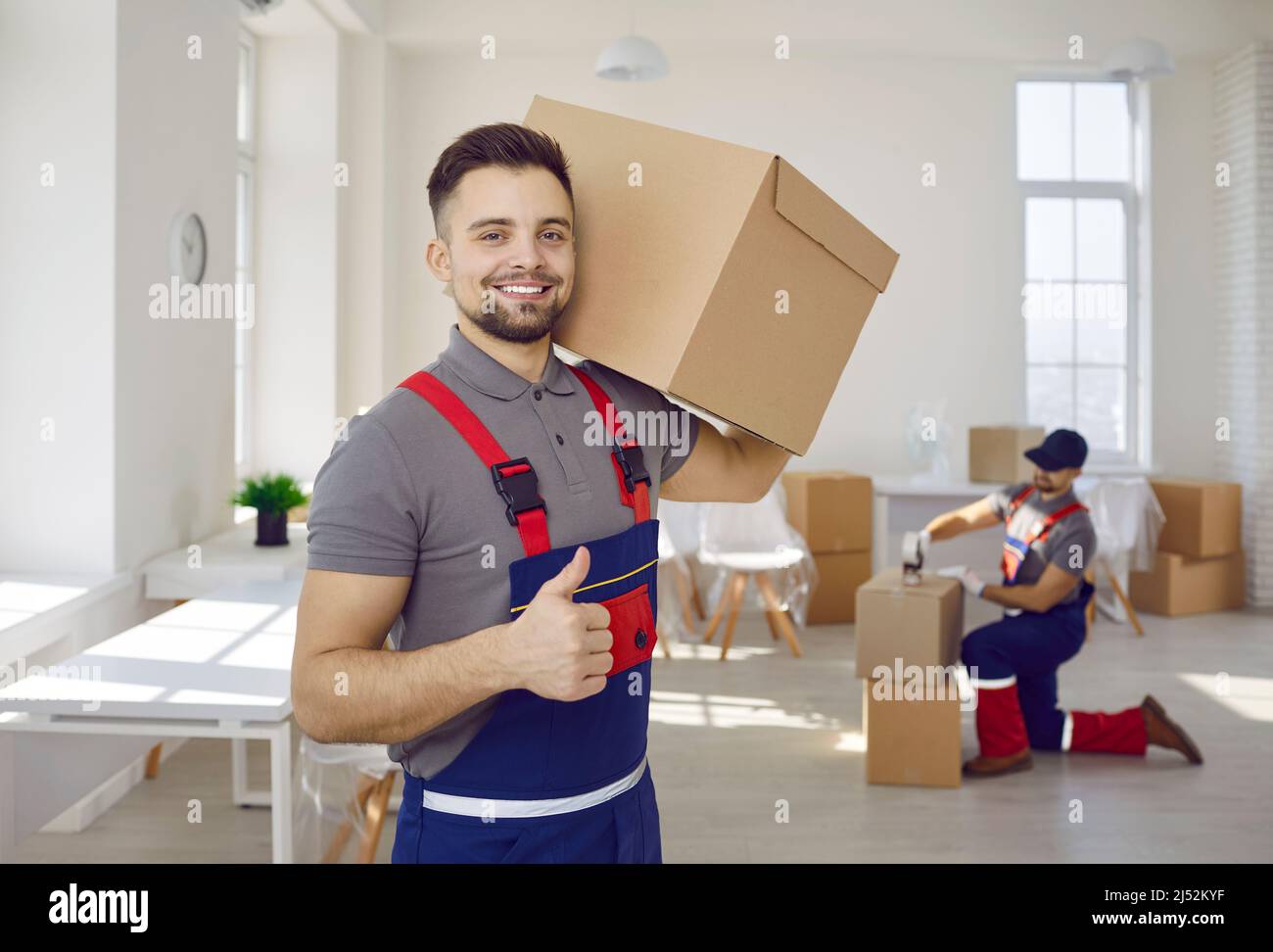 Lächelnder männlicher Arbeiter des Umzugsunternehmens und des Zustellers, der einen Karton mit Daumen nach oben hält. Stockfoto