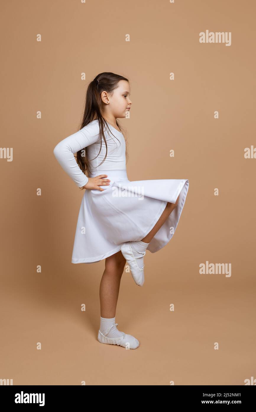 Seitenansicht eines jungen niedlichen ernsthaften Mädchens mit langen dunklen Haaren in weißem Kleid, Socken und Turnschuhen stehen, die Hände an der Taille halten und das Bein hochheben Stockfoto