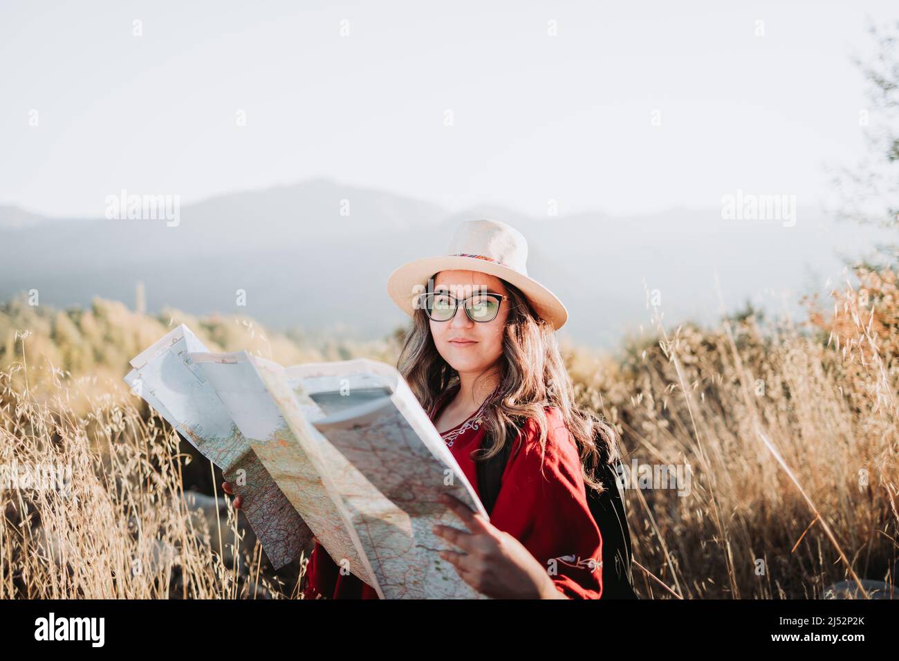 Junge Frau, die einen Hut, einen roten Poncho und einen Rucksack trägt, überprüft die Karte während der Wanderung, um die Natur zu erkunden. Stockfoto