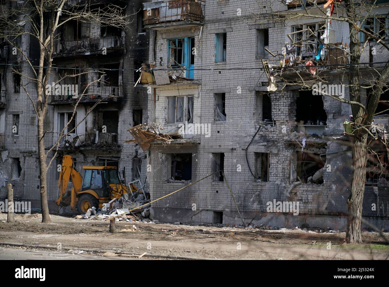 Ein verbranntes, zerstörtes Wohnhaus ohne Fenster, das während des Beschuss und der Militäroperationen in der Ukraine beschädigt wurde. Unter dem Haus räumt der Traktor Stockfoto